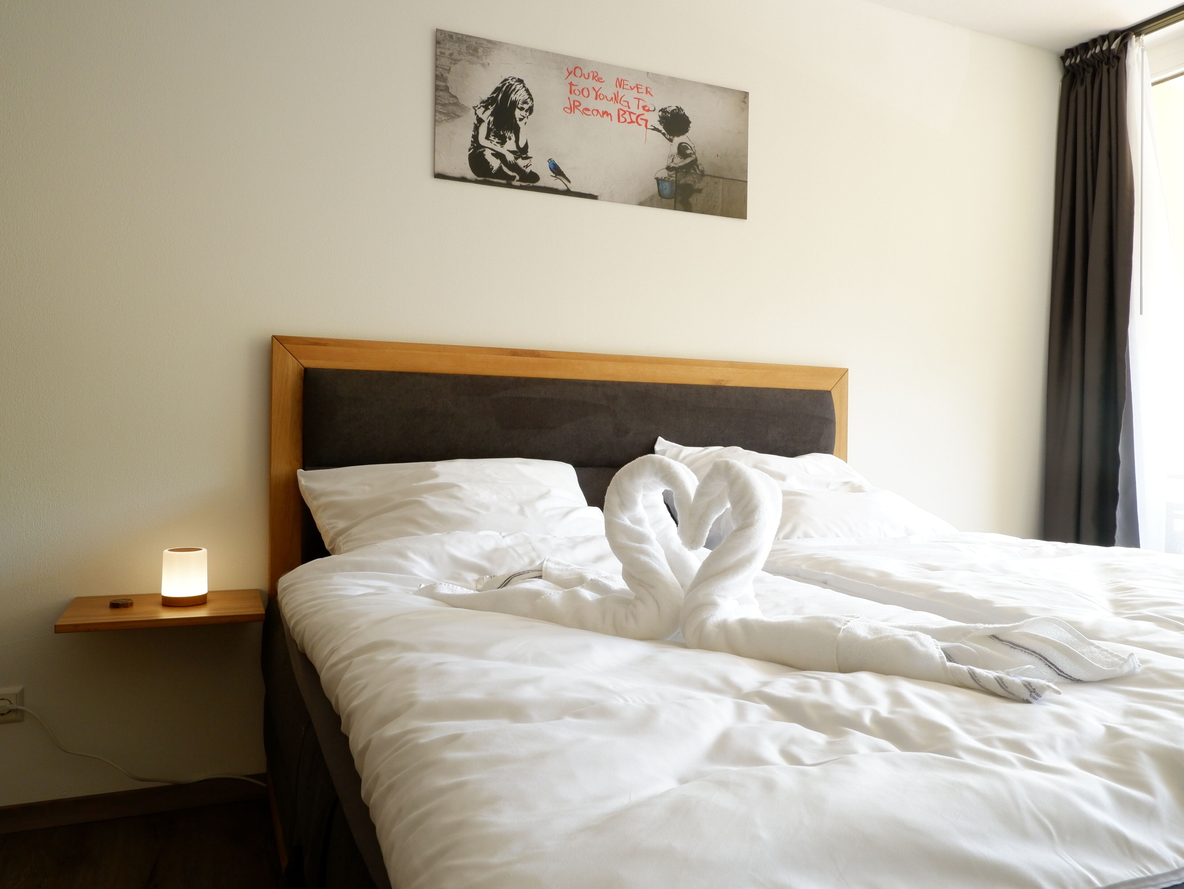 Un lit bien fait, avec du linge de lit blanc et une disposition de serviettes en forme de cœur, une table de chevet avec une lampe et une œuvre d’art accrochée au mur.