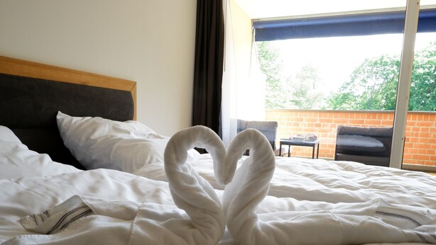 A hotel room with a bed, a heart-shaped towel arrangement, a window with a view of trees, and a brick wall. - Room 116 at the AlbThermen (Bad Urach)