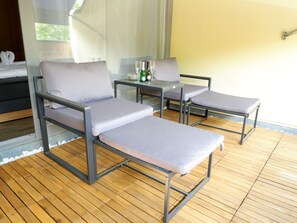 Een zonnig terras met metalen stoelen en een tafel, met uitzicht op groen door een raam.