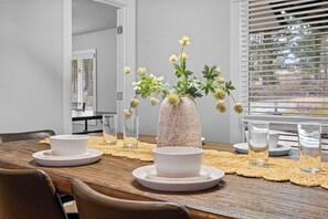 Ein Esstisch mit weißen Schalen und Gläsern, eine Vase mit Blumen und ein Fenster mit Jalousien.