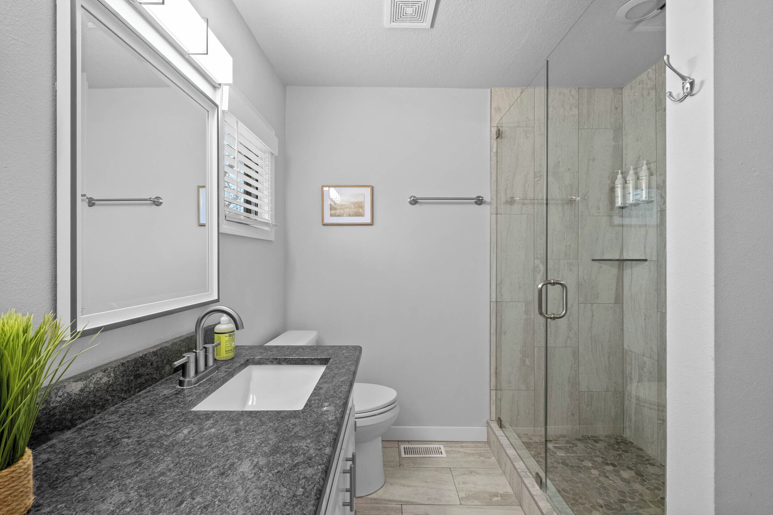 A bathroom with a granite countertop, a white sink, a toilet, a framed picture on the wall, and a glass-enclosed shower area.
