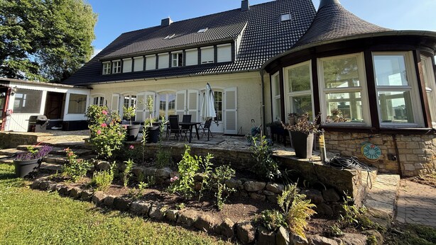 Ett tvåvåningshus med ett tak täckt av takpannor, en trädgård med olika växter och en uteplats med utemöbler.