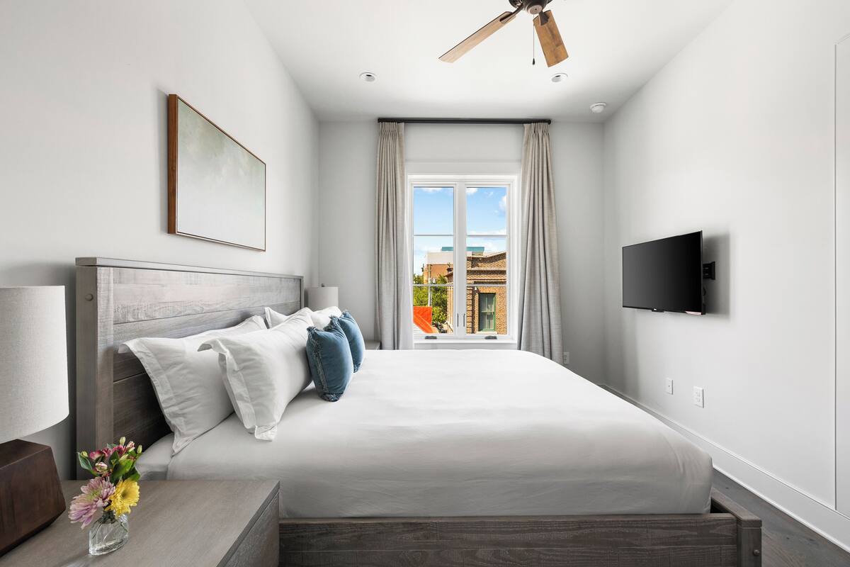 A modern hotel room with a large bed, a ceiling fan, a TV, and a window with a view of buildings.