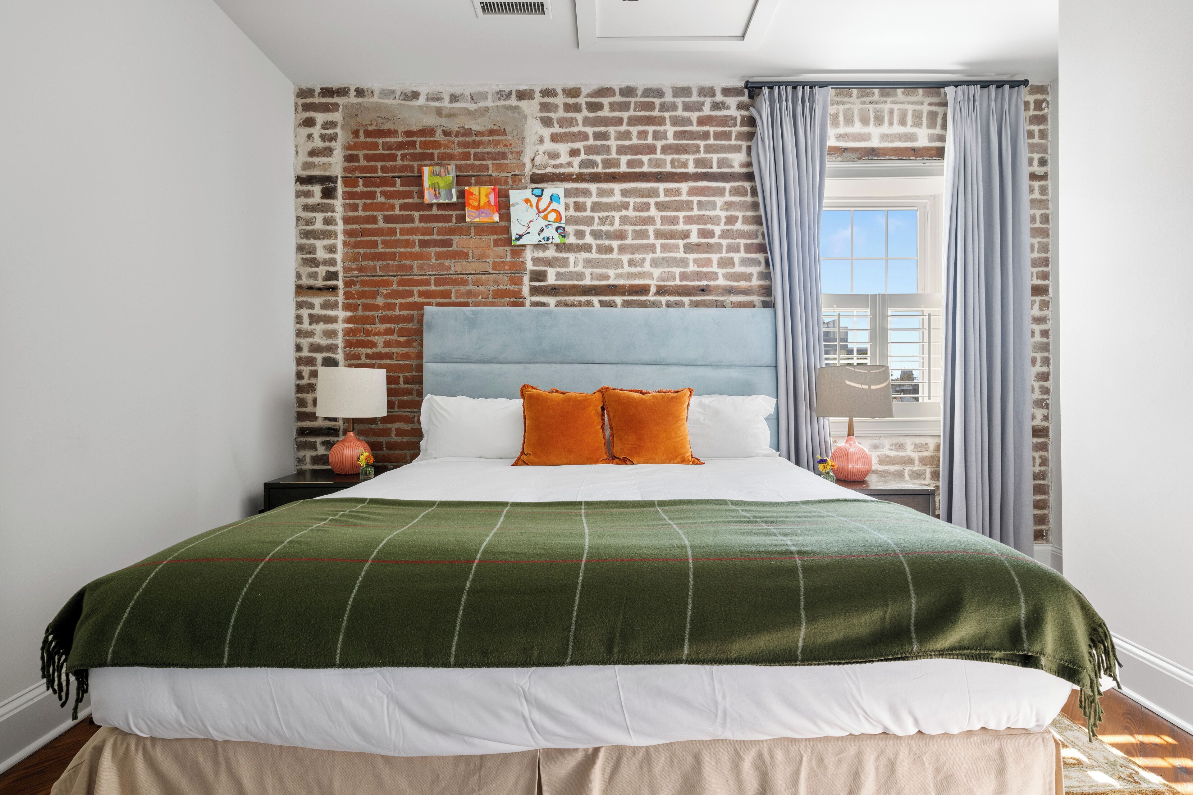 Habitación de hotel con pared de ladrillo, cama con ropa de cama blanca y almohadas naranjas, una manta verde, una mesita de noche con lámpara y vistas al exterior a través de una ventana con cortinas.