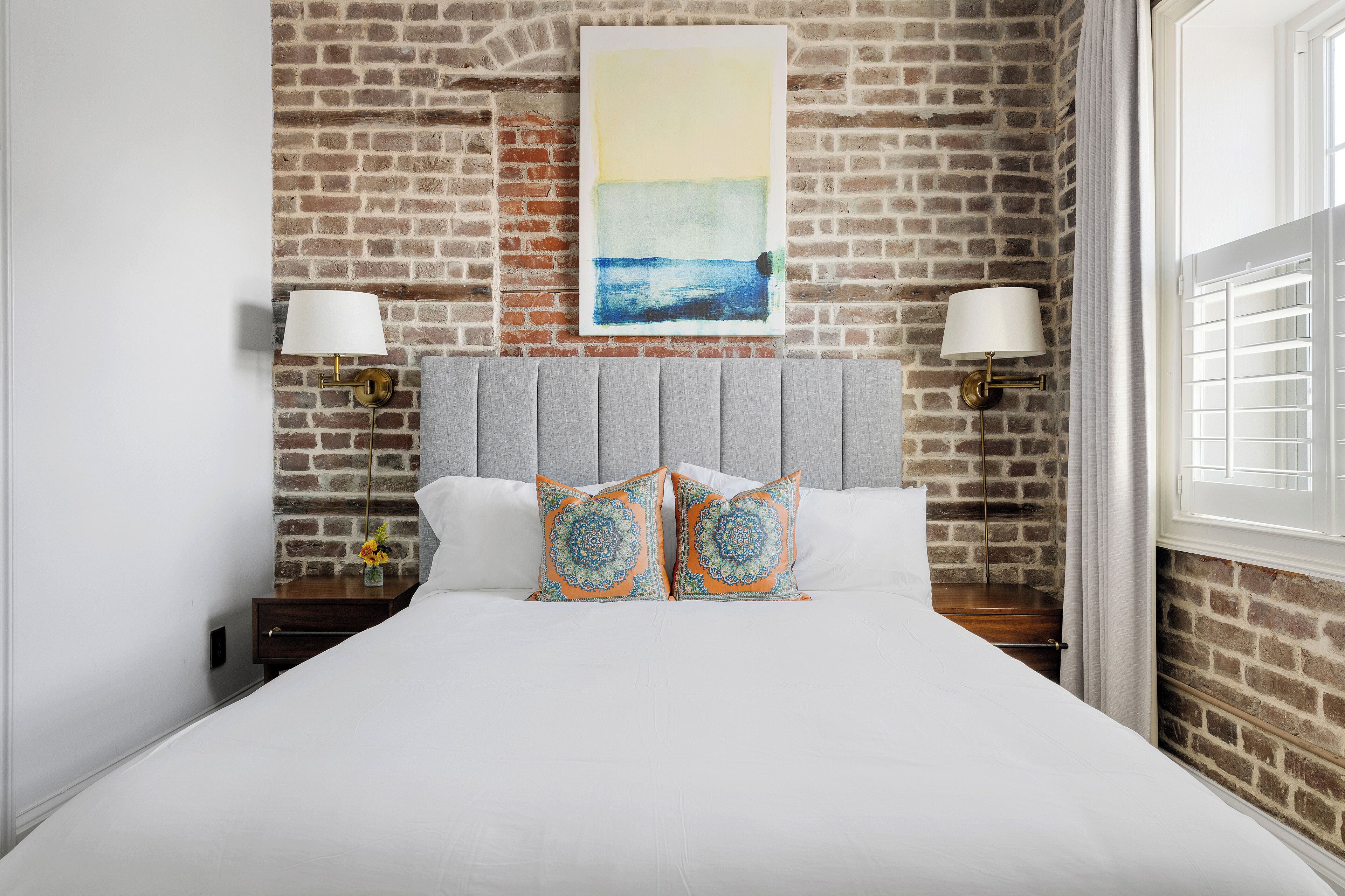 Quarto de hotel com parede de tijolos, uma cama com roupa de cama branca, duas almofadas decorativas, uma mesa de cabeceira com abajur e um quadro sobre a cama.