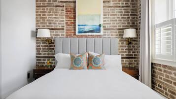 Quarto de hotel com parede de tijolos, uma cama com roupa de cama branca, duas almofadas decorativas, uma mesa de cabeceira com abajur e um quadro sobre a cama.