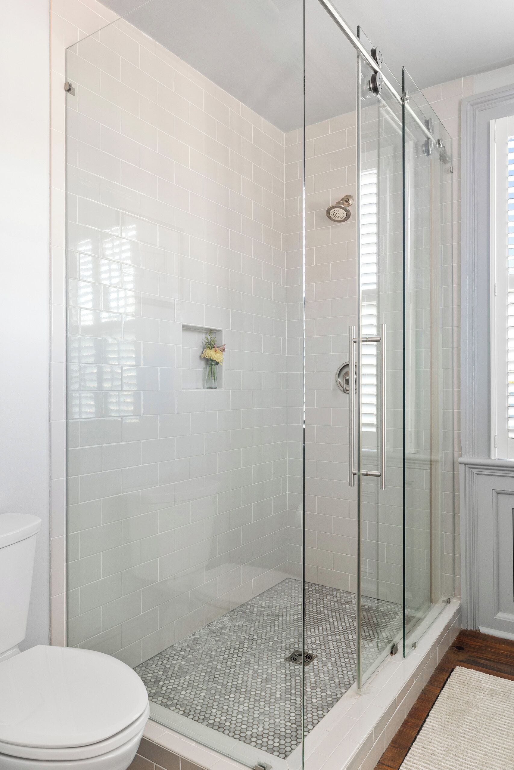 Une douche fermée par des parois de verre, avec des murs recouverts de carreaux blancs et des toilettes blanches.