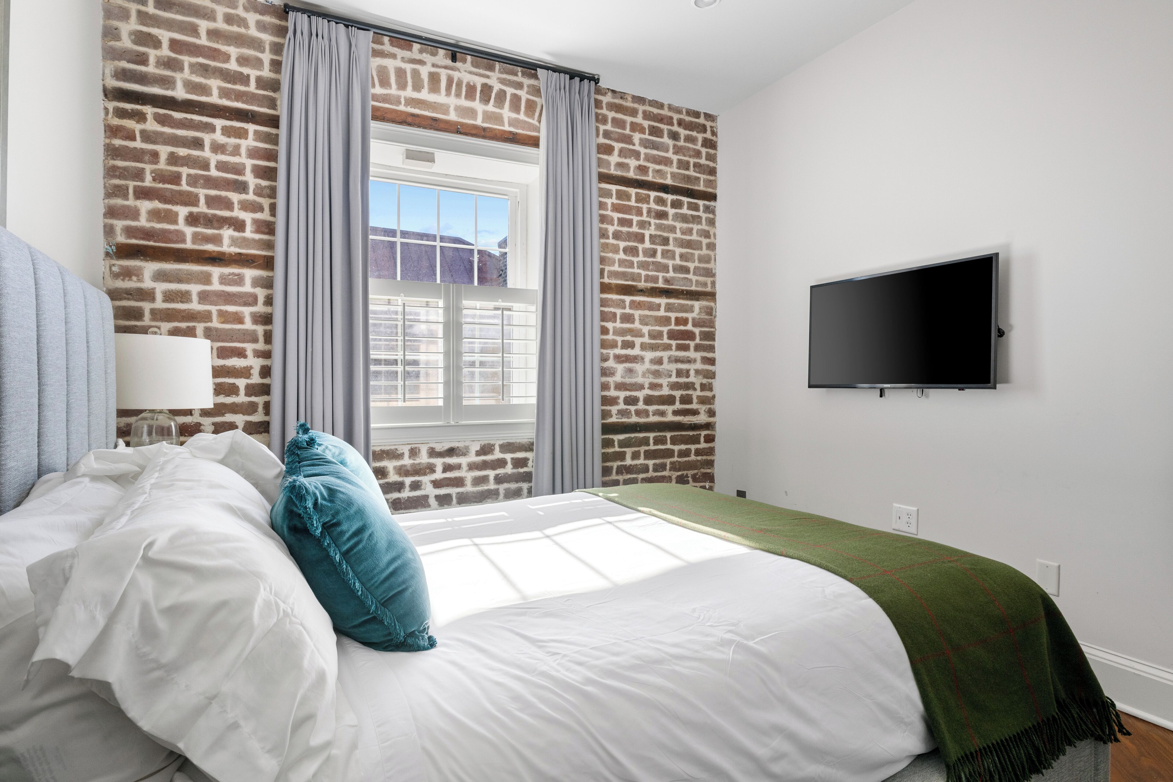 A bedroom with a brick wall, a window with curtains, a bed with white and teal pillows, and a flat-screen TV mounted on the wall.