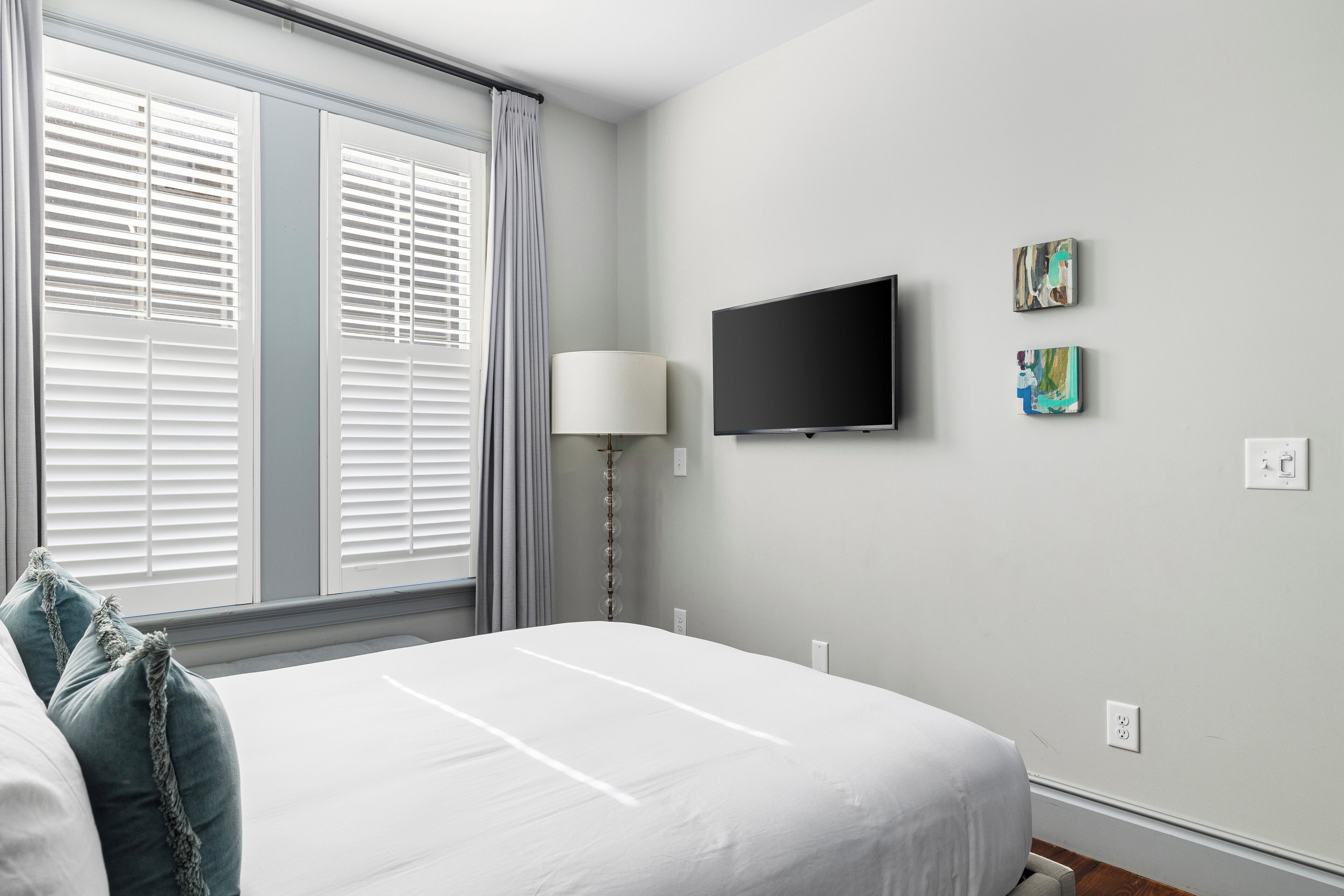 Quarto de hotel com cama, televisão presa na parede, janela com persianas e luminária.
