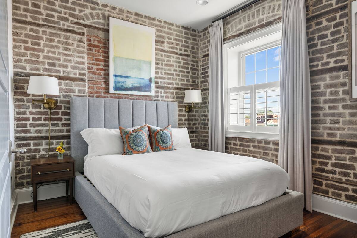 A bedroom with a brick wall, a large bed with grey bedding, two bedside lamps, and a window with curtains.