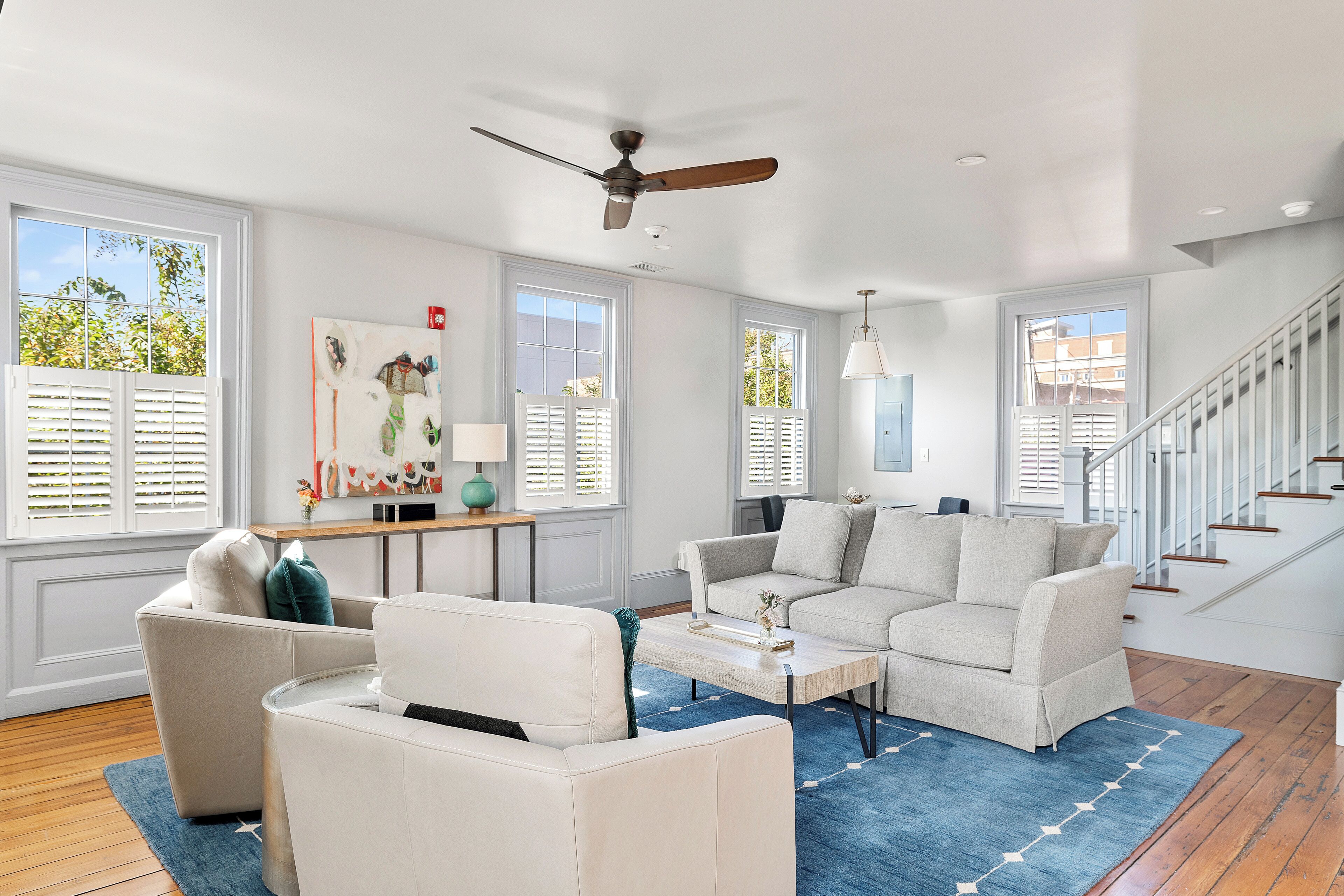 A spacious living room with a ceiling fan, large windows with blinds, a staircase, and a blue rug.