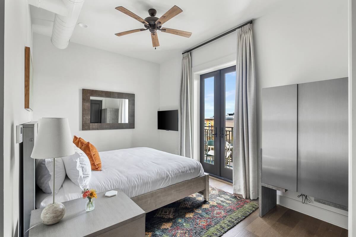 A modern bedroom with a bed, a TV, a ceiling fan, a window with curtains, and a balcony view.