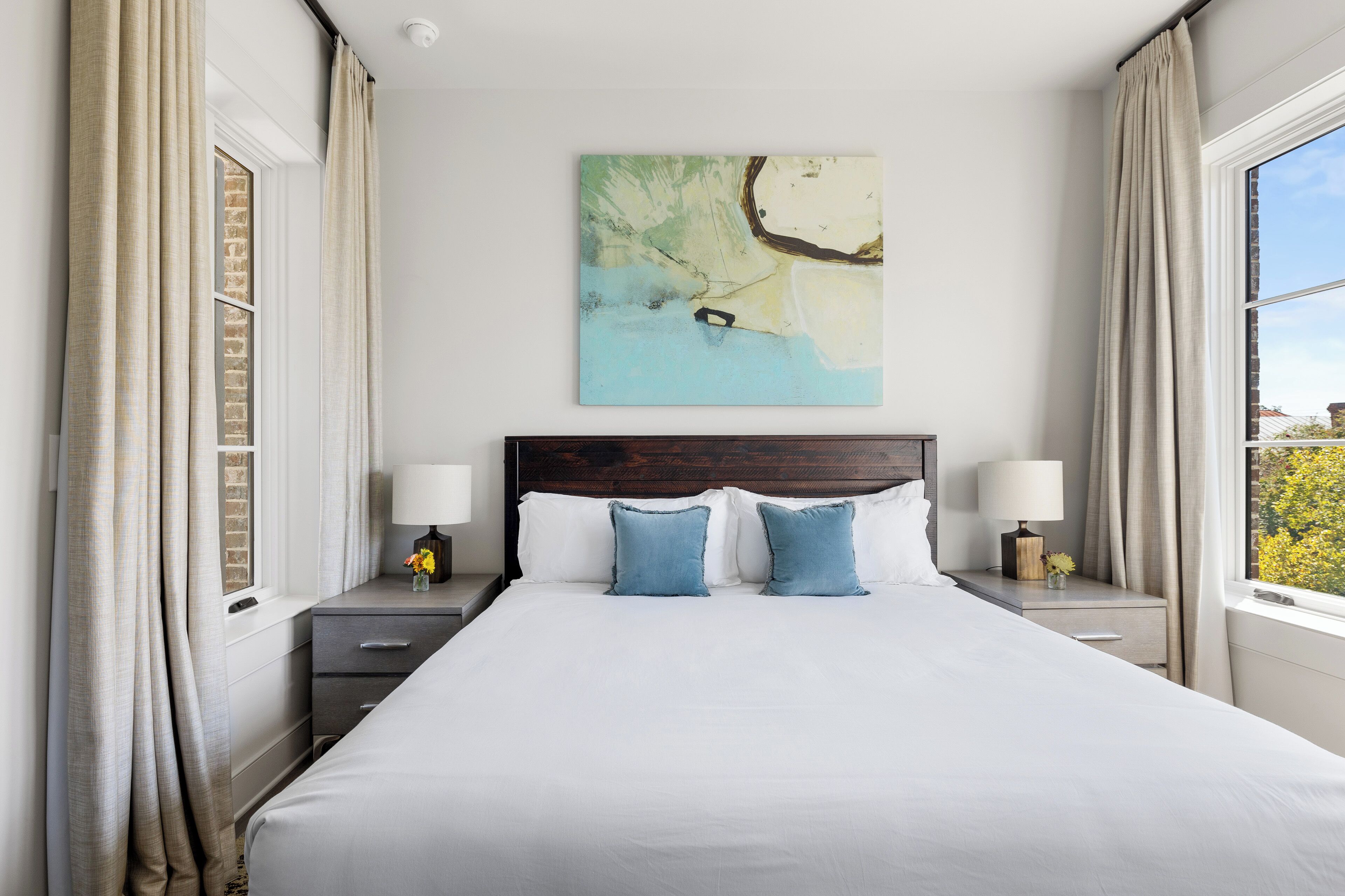 Een hotelkamer met een groot bed, twee nachtkastjes met lampen, een kunstwerk aan de muur en een raam met gordijnen.