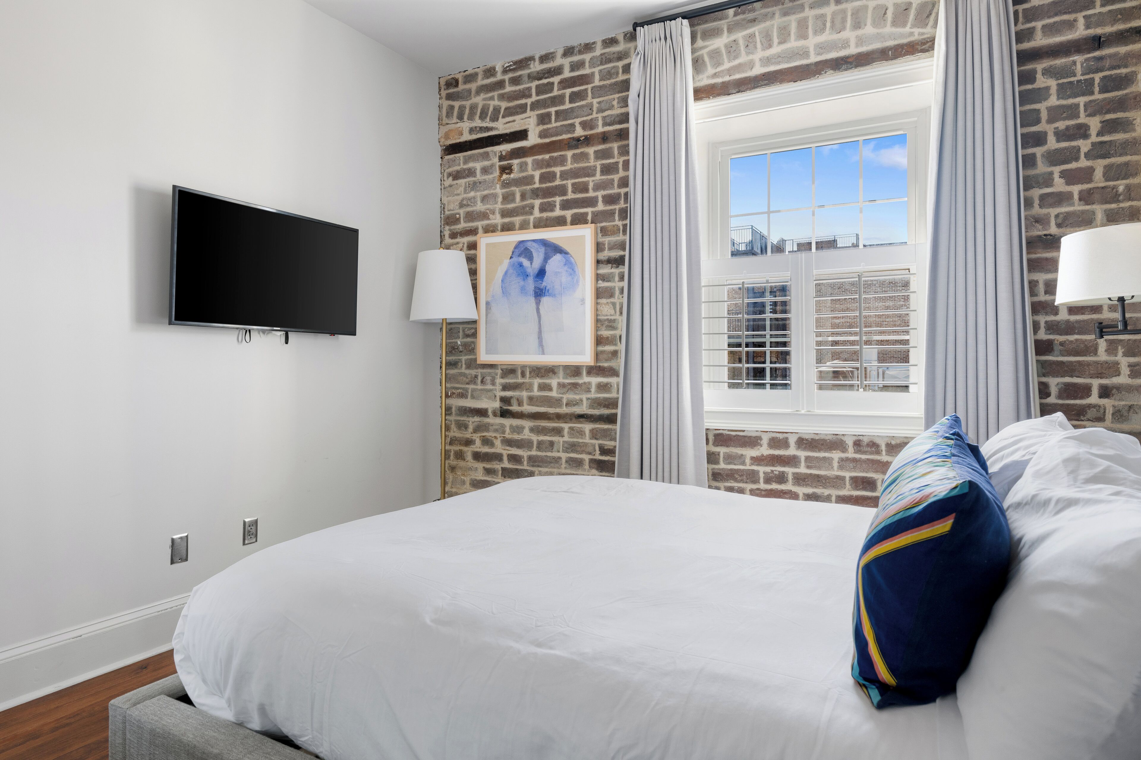 A bedroom with a brick wall, a flat-screen TV, a window with curtains, and a bed with white bedding.