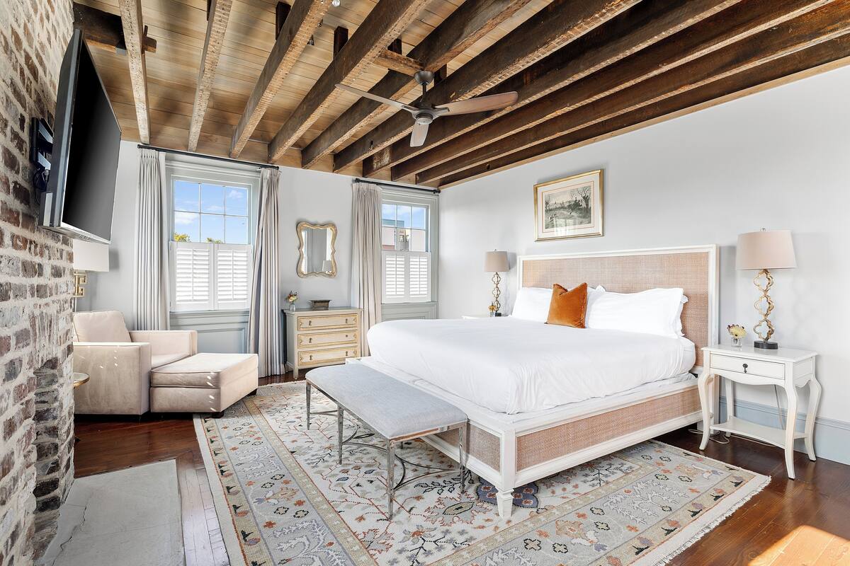 A bedroom with a large bed, a bench, a dresser, a nightstand, and a TV mounted on a brick wall.
