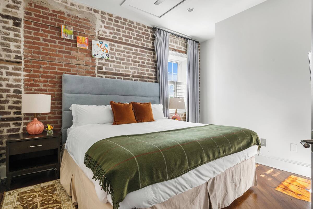 A bedroom with a brick wall, a bed with a green blanket, a nightstand with a lamp, and a window with curtains.