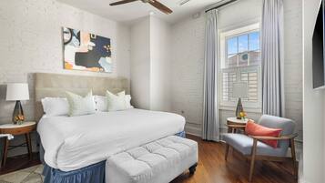 Quarto de hotel com uma cama grande, uma cadeira, um ventilador de teto, uma janela com cortinas e um quadro na parede.