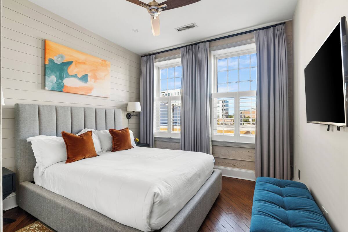 A bedroom with a bed, a bench, a ceiling fan, a TV, and a window with curtains.