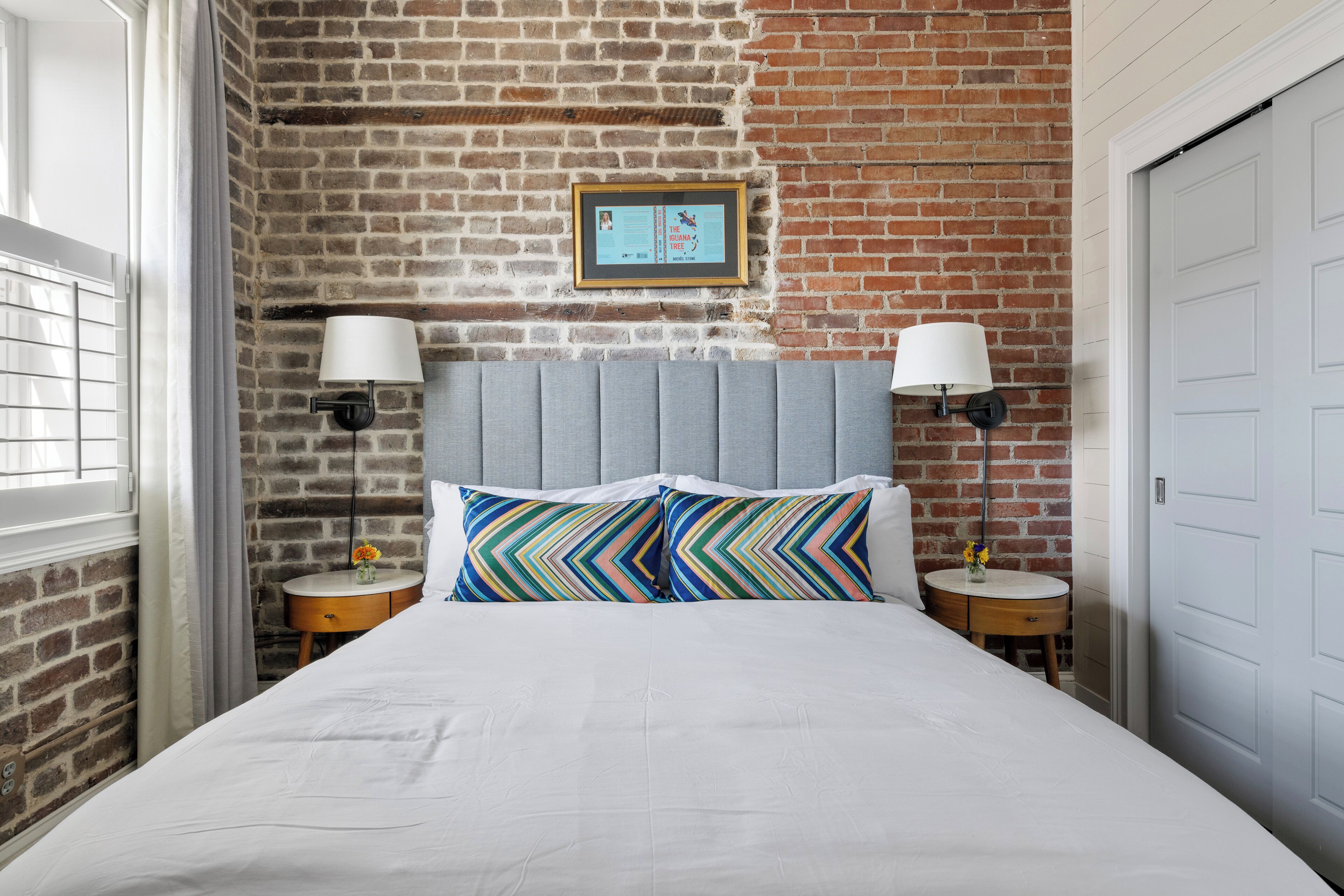 A hotel room with a brick wall, a bed with white bedding, two nightstands with lamps, and a framed picture on the wall.