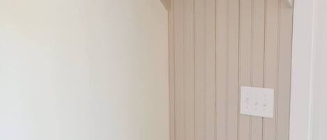 Un banco blanco con respaldo de listones y un estante encima, fijado a una pared con paneles verticales.
