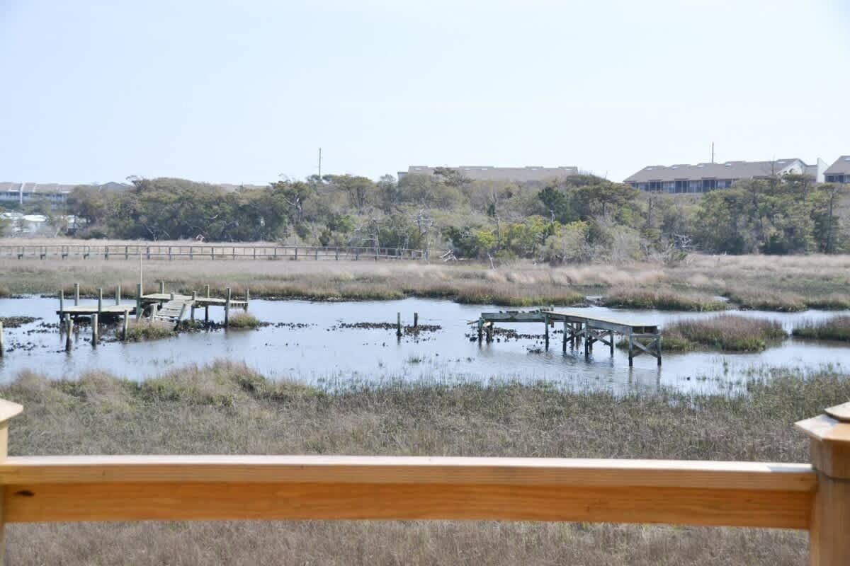A view of a wetland area with wooden docks extending into a body of water, surrounded by sparse vegetation and distant buildings.