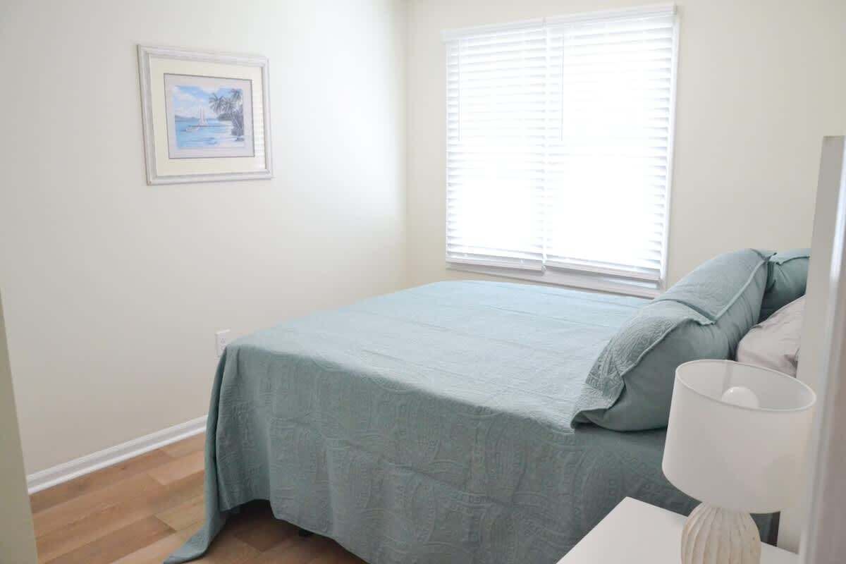A bedroom with a bed, a nightstand, a lamp, a window with blinds, and a framed picture on the wall.