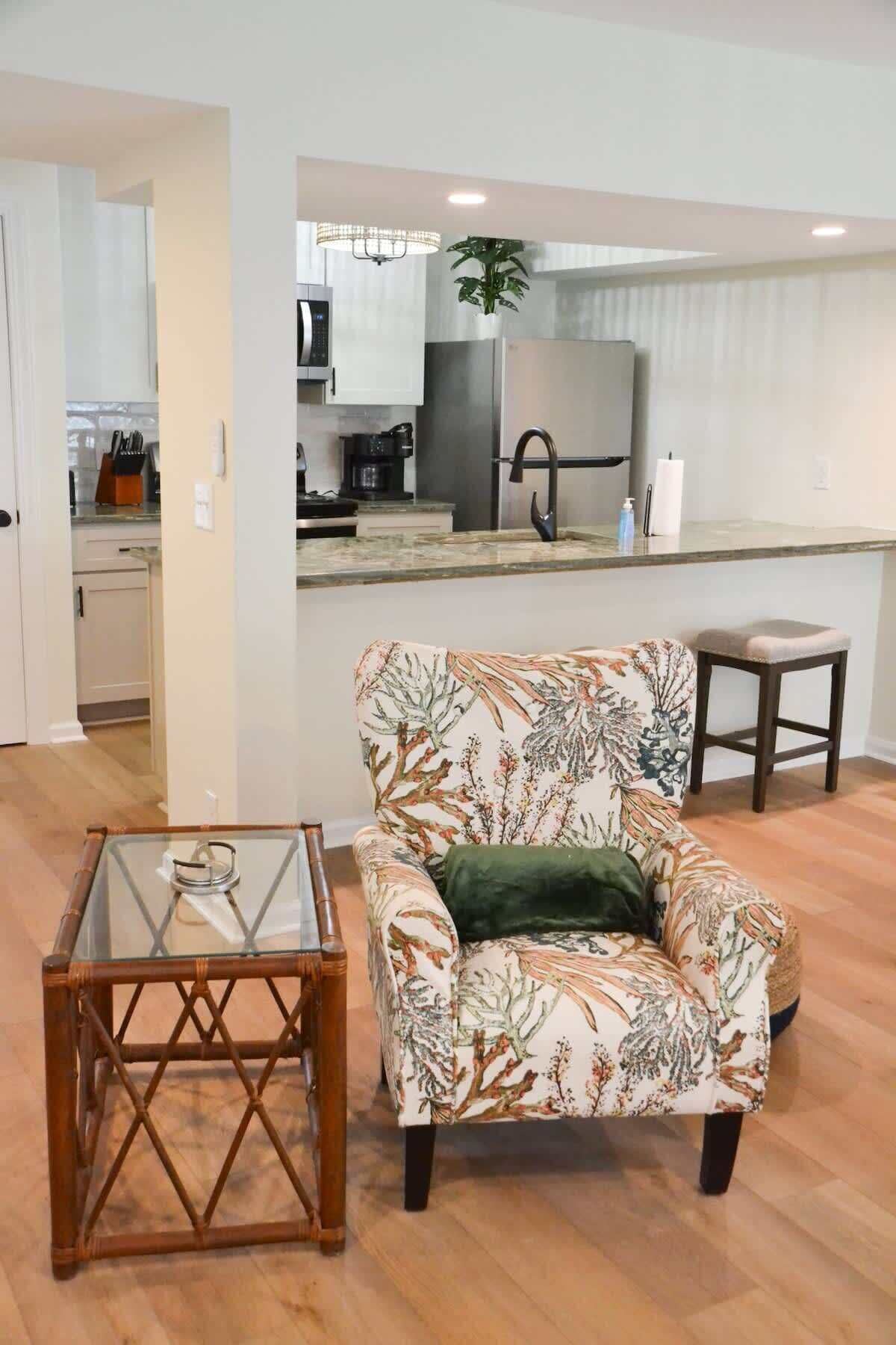 A living room with a floral armchair, a glass-top coffee table, and a wooden stool.