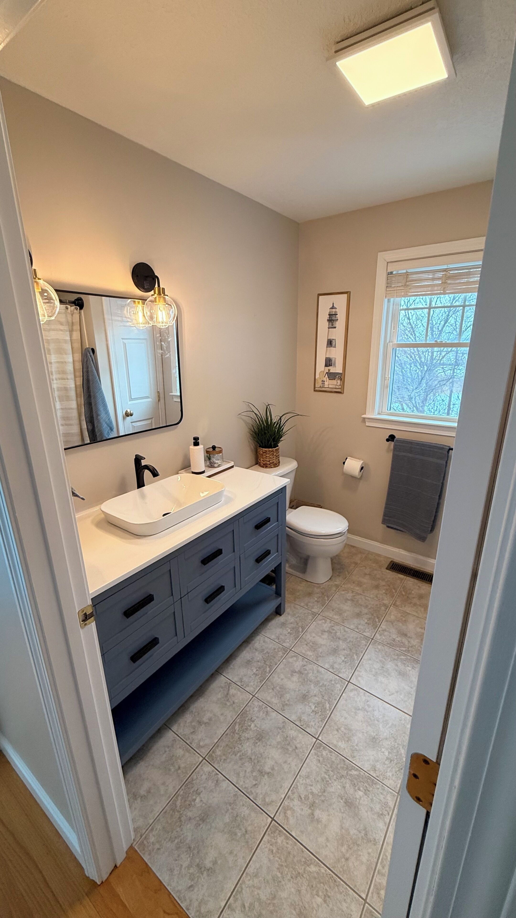 A bathroom with a white sink, dark blue cabinet, and a toilet. There is a window with blinds and a plant on the toilet.