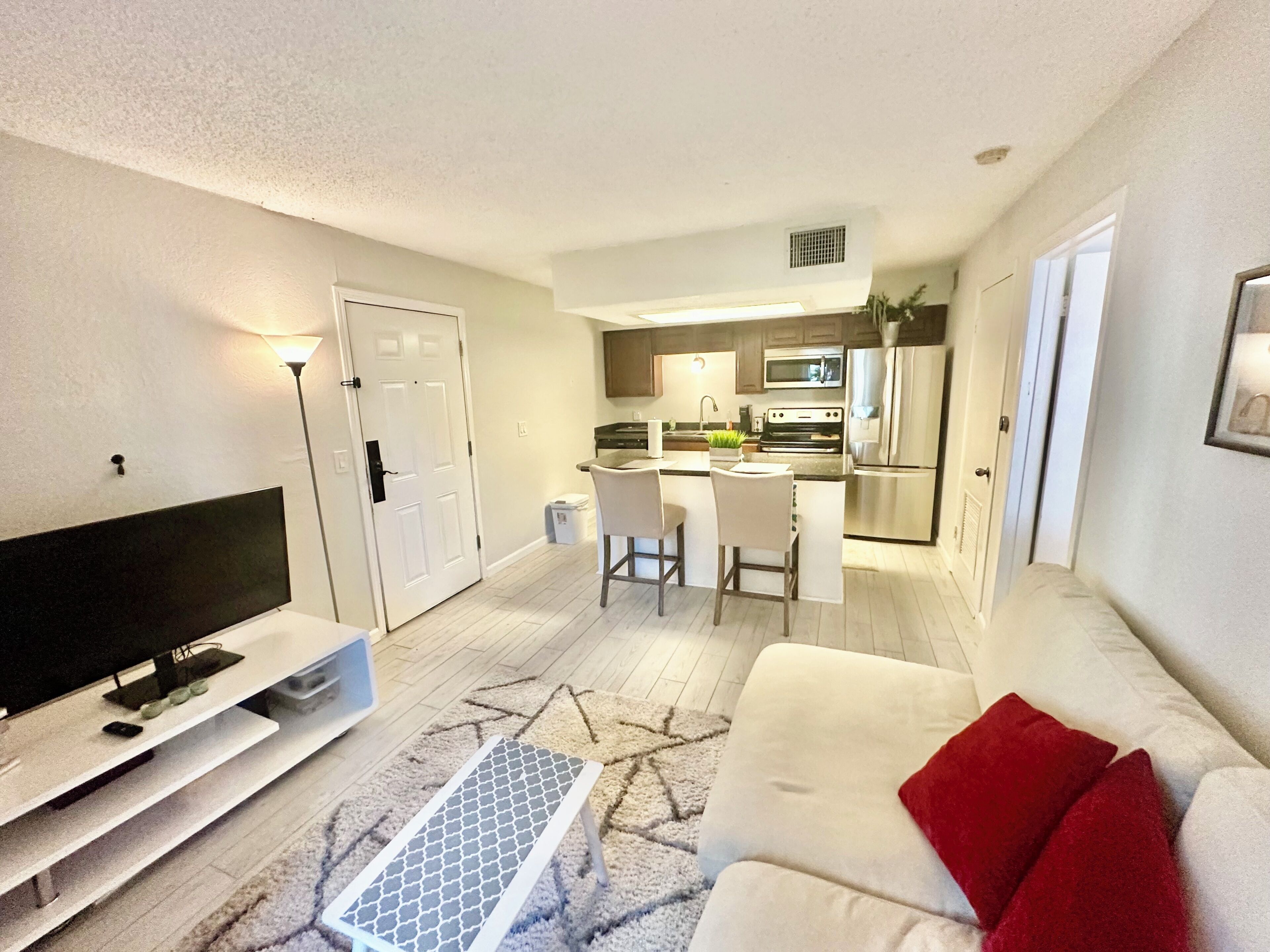 A modern living room with a flat-screen TV, a white sofa with red cushions, a patterned rug, and a kitchen in the background.