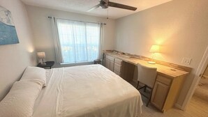 Een slaapkamer met een bed, een bureau, een stoel, een plafondventilator, een raam met gordijnen en een lamp.