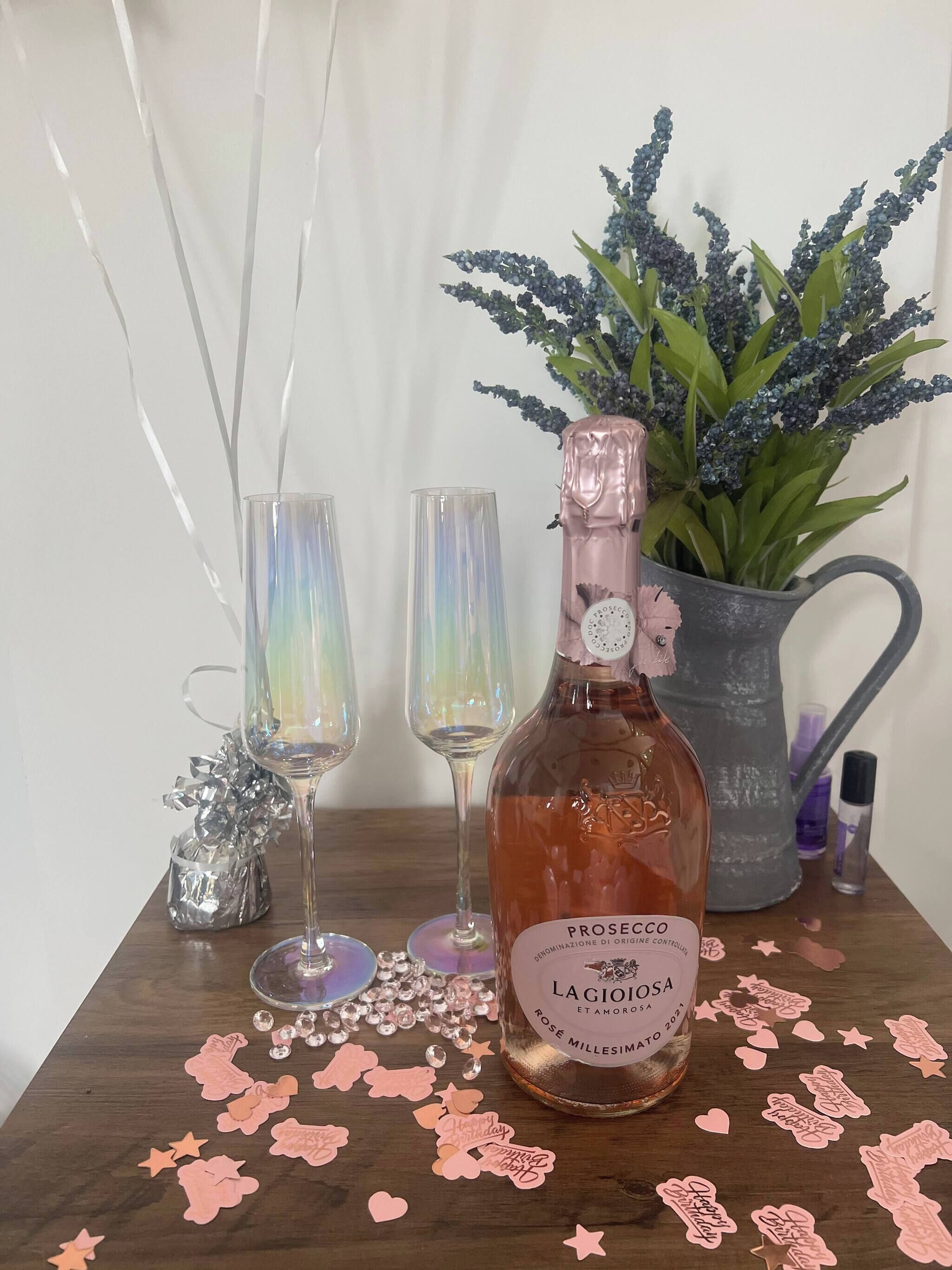 A bottle of Prosecco, two glasses, a decorative plant, and scattered confetti on a wooden surface.