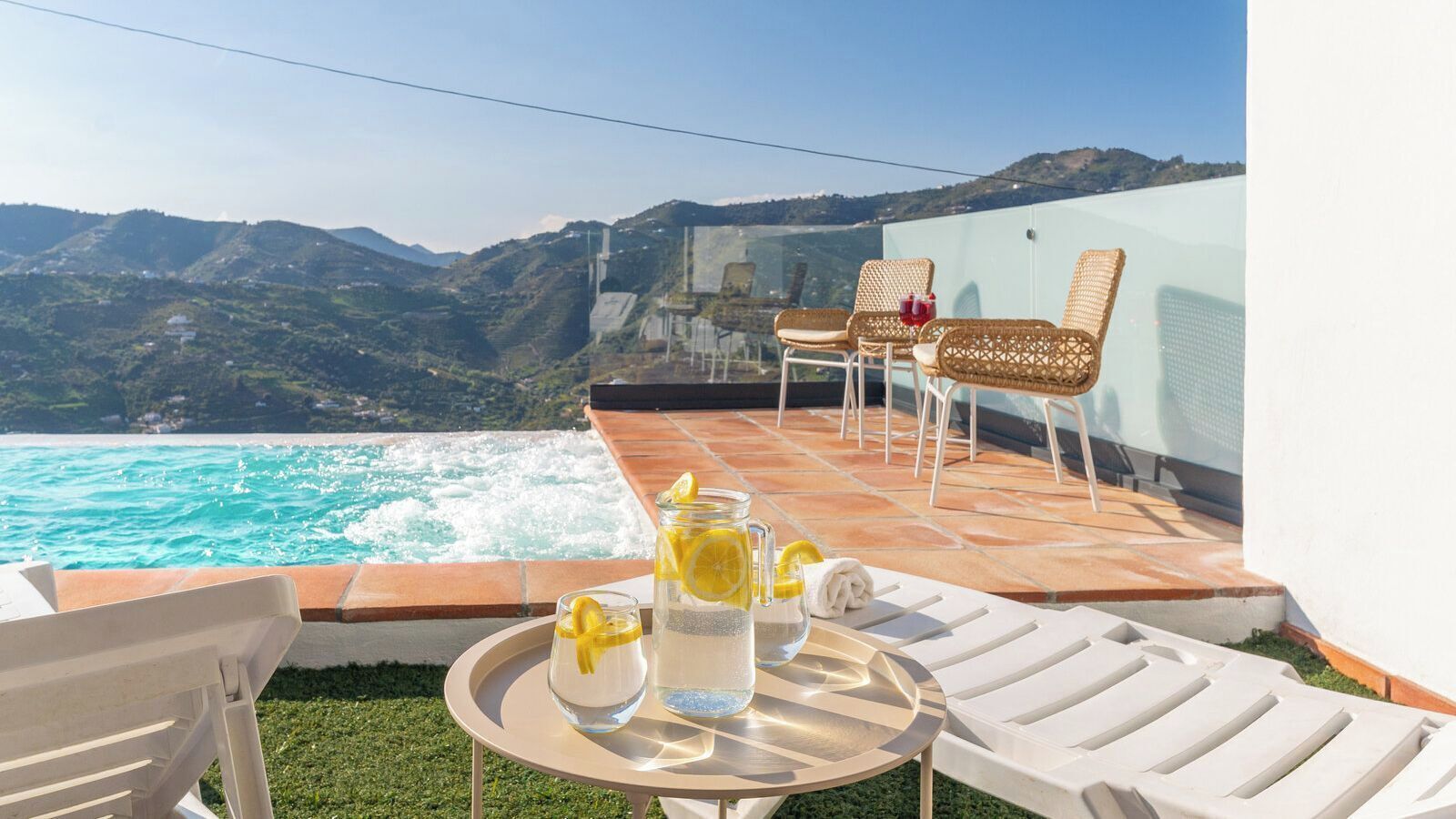 Une terrasse avec une piscine, des fauteuils en osier, une table avec des boissons et un paysage montagneux en toile de fond.