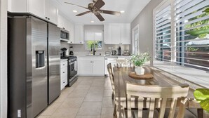 A modern kitchen with white cabinetry, stainless steel appliances, a dining area with a wooden table, and a view through large windows. - 10 Min to Mission Beach & Sea World | Kids Playset (San Diego)