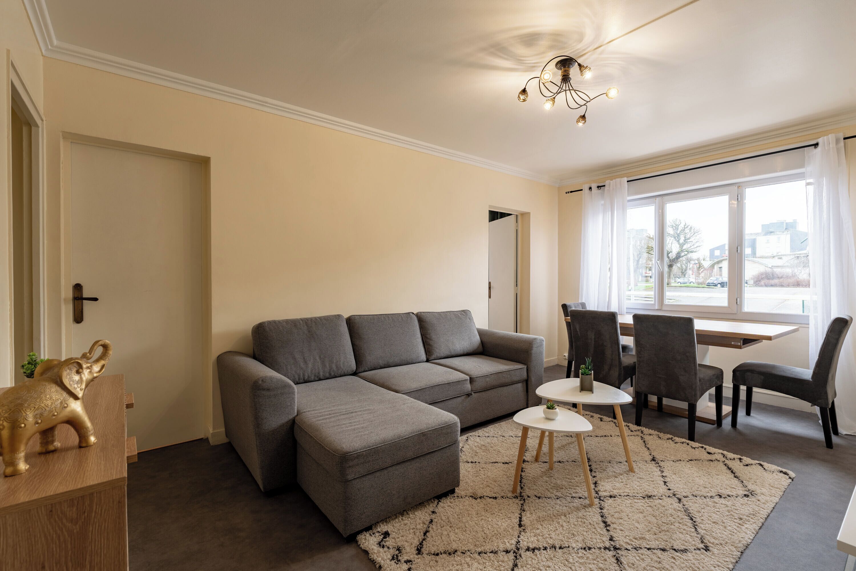 A living room with a grey sectional sofa, a small white table, and two chairs. There is a wooden cabinet with a decorative elephant figurine.