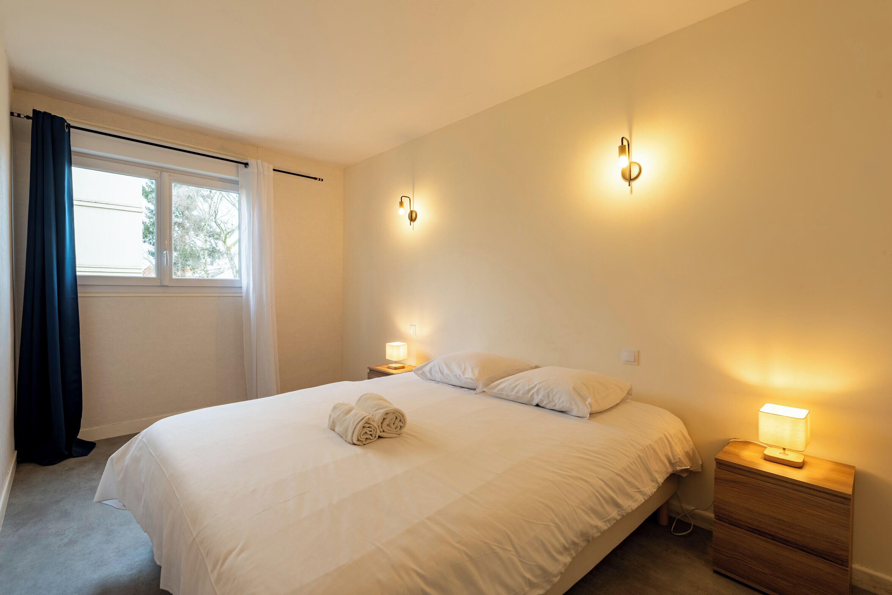 Un dormitorio con una cama, dos lámparas de noche, una ventana con cortinas y una toalla enrollada sobre la cama.