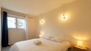 Quarto com cama, duas luminárias de cabeceira, janela com cortinas e uma toalha enrolada na cama.