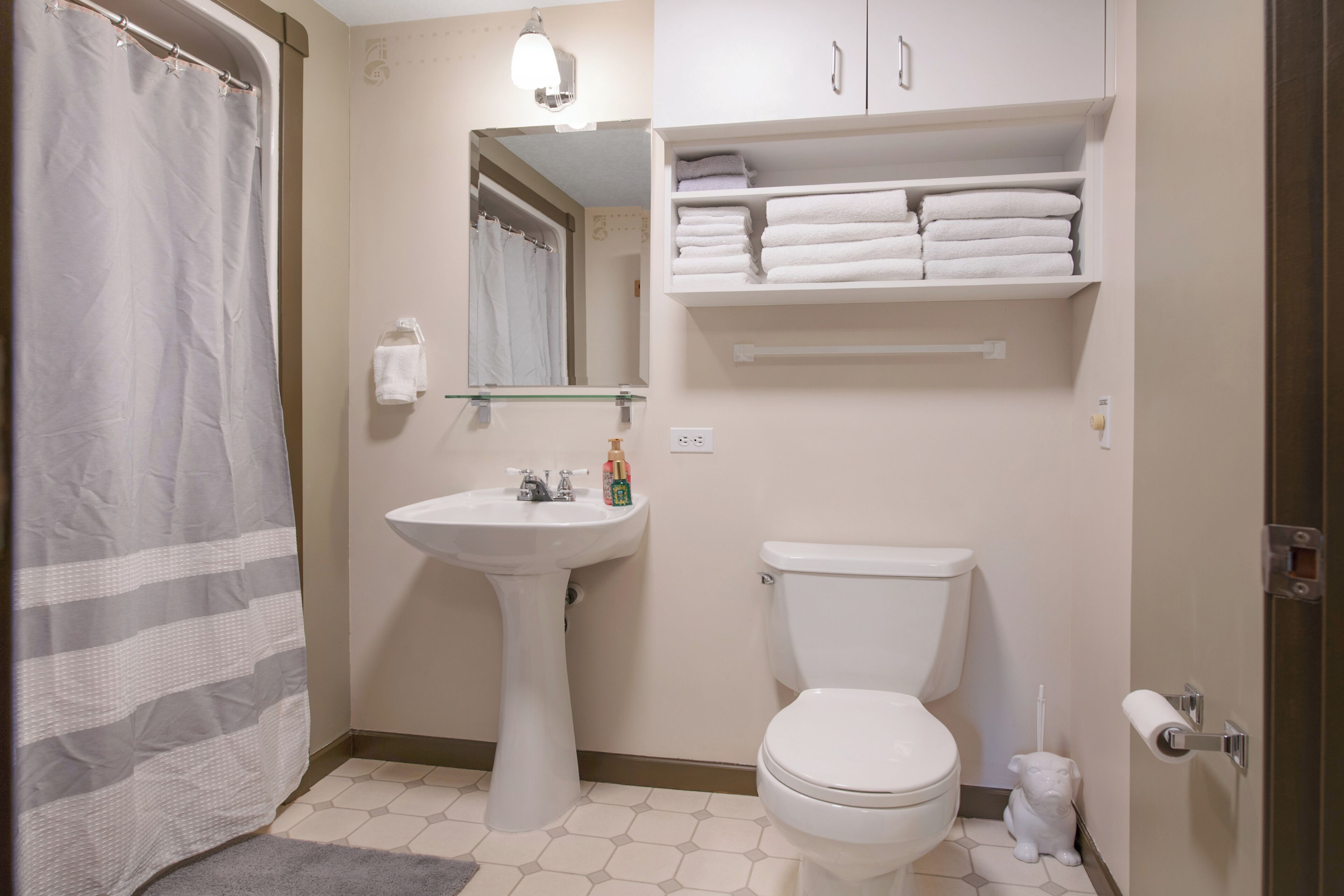 A bathroom with a shower curtain, a pedestal sink, a toilet, a mirror, and a shelf with towels.