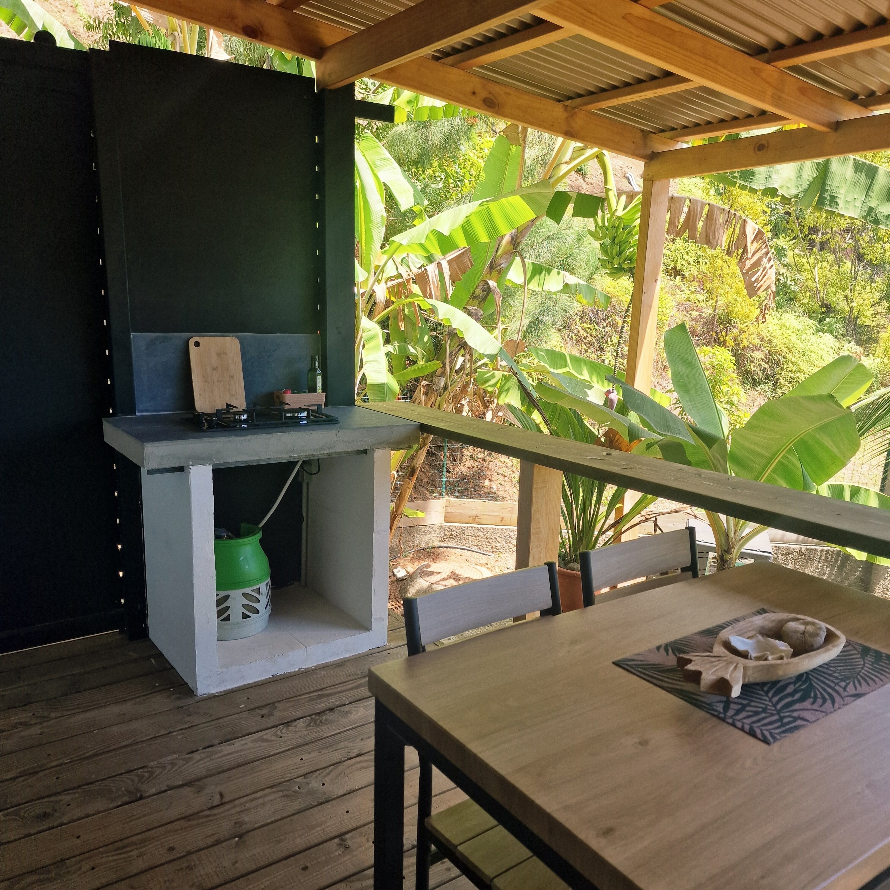 An outdoor dining area with a wooden table and chairs, a grill, and a view of lush greenery.