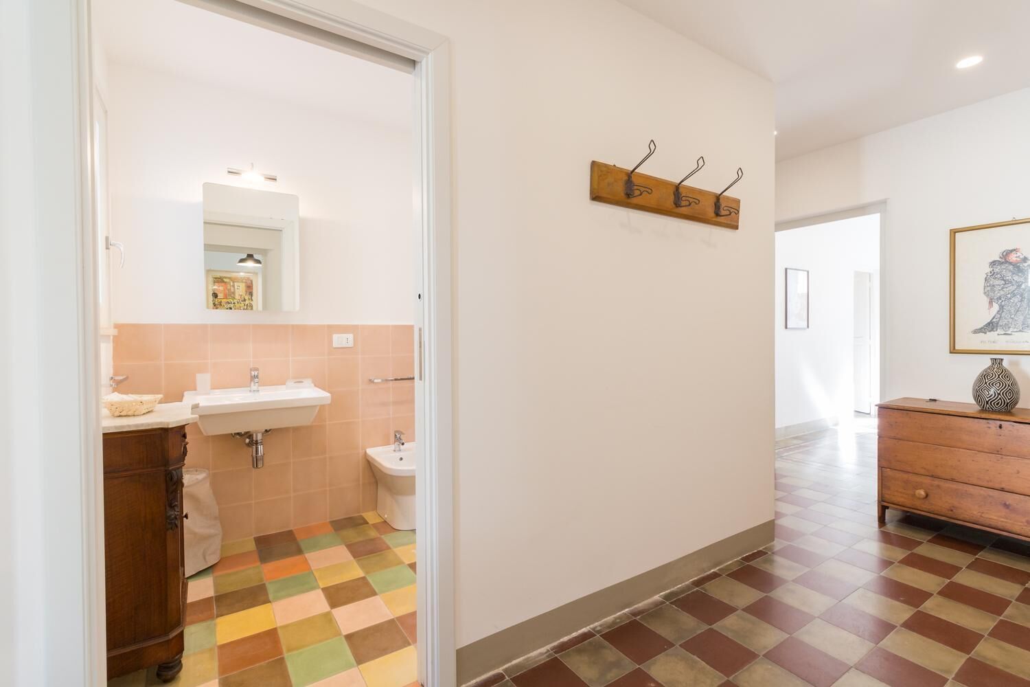 Corredor com banheiro, contendo pia, vaso sanitário e espelho. Há uma cômoda de madeira e um quadro na parede. O chão possui um padrão quadriculado.