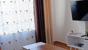 Ein Wohnzimmer mit einer Couch, einem Couchtisch, einem Fernseher auf einem Schrank und einem Fenster mit Vorhängen.