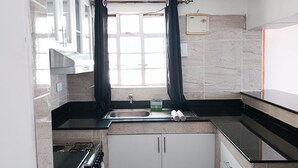 Eine kompakte Küche mit Spüle, Herd und Schränken, ein Fenster mit Vorhängen sowie eine schwarze Matte mit Besteck auf dem Boden.