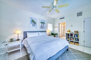 Une chambre à coucher comprenant un lit, des tables de chevet, un ventilateur de plafond, une bibliothèque et une porte donnant sur une autre pièce.