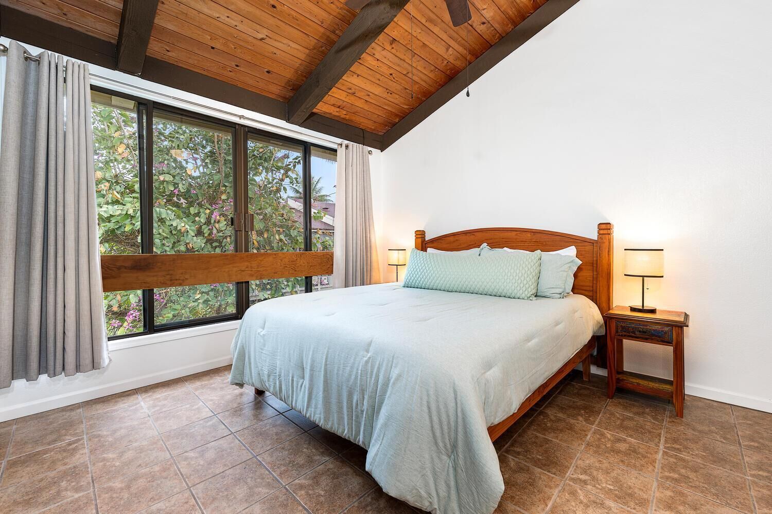 A bedroom with a large window, wooden ceiling beams, a bed with a headboard, and two bedside tables with lamps.