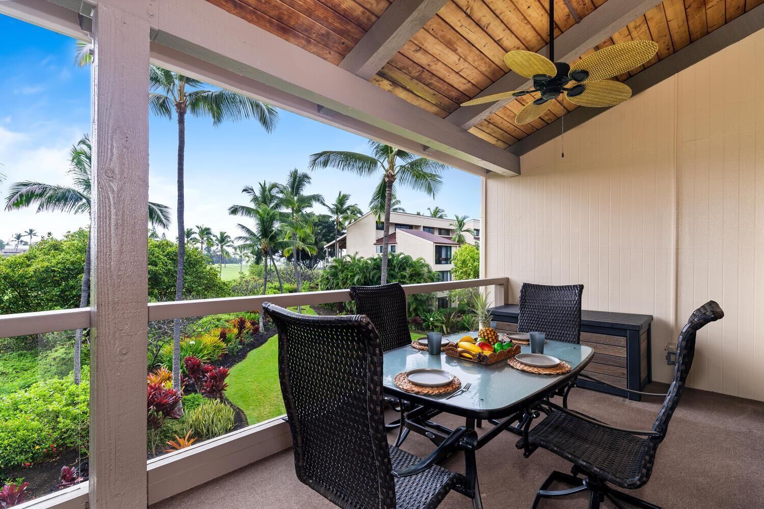 A covered outdoor dining area with wicker furniture, a table set with plates and a fruit basket, and a view of palm trees and a building in the background.