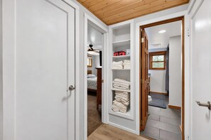 A hallway with a wooden ceiling, white walls, and a door leading to a bedroom with a bed and a closet with folded towels.