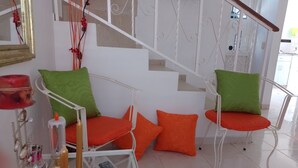 Un saló modern amb una escala, dues cadires de metall blanc amb coixins verds i taronja, un comandament sobre una taula de vidre i un objecte decoratiu a la paret.