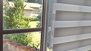Finestra amb persianes horitzontals, vista a vegetació a l'exterior i una part d'un teixit estampat.