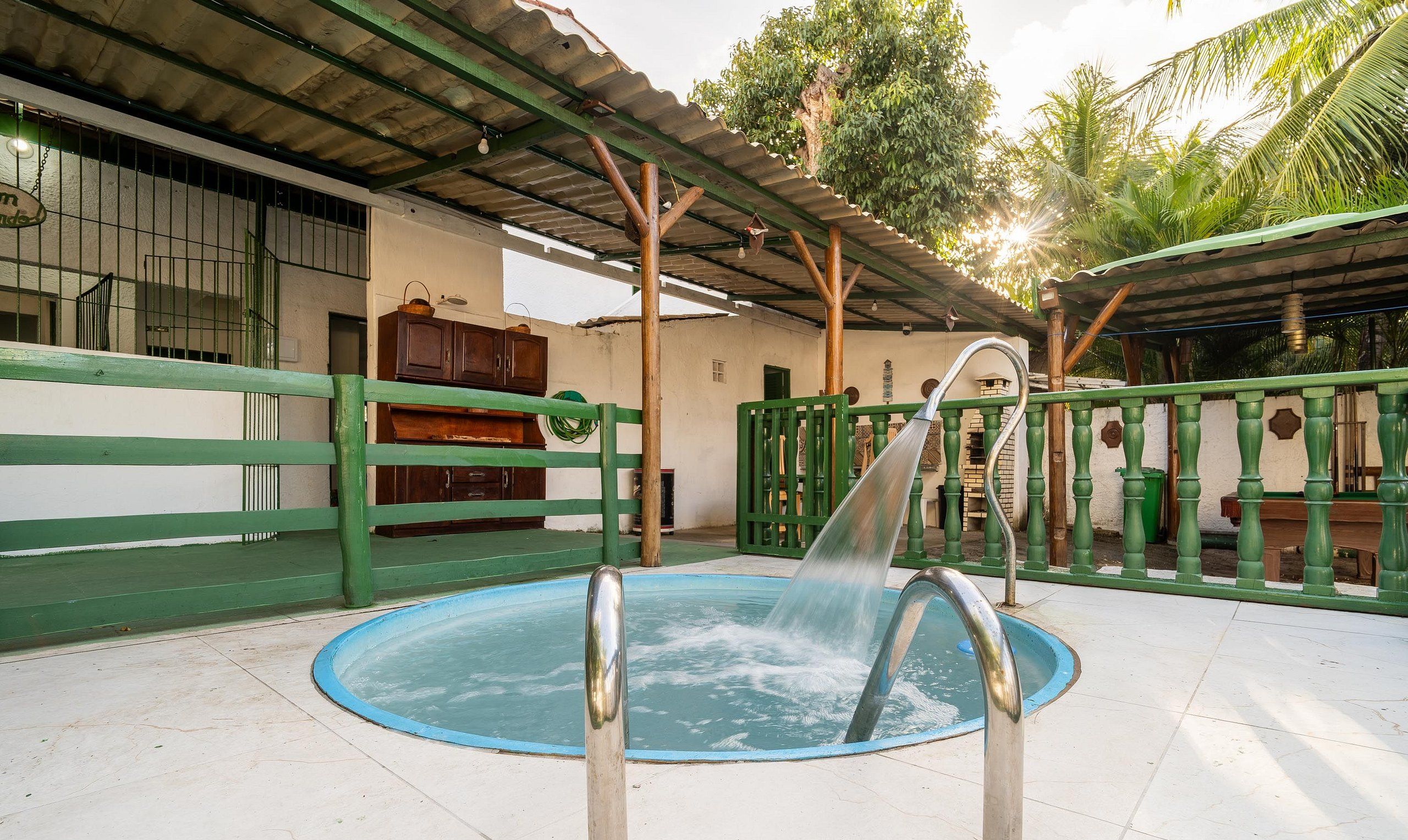 A swimming pool with a water fountain, surrounded by a green fence and a covered area with a roof.