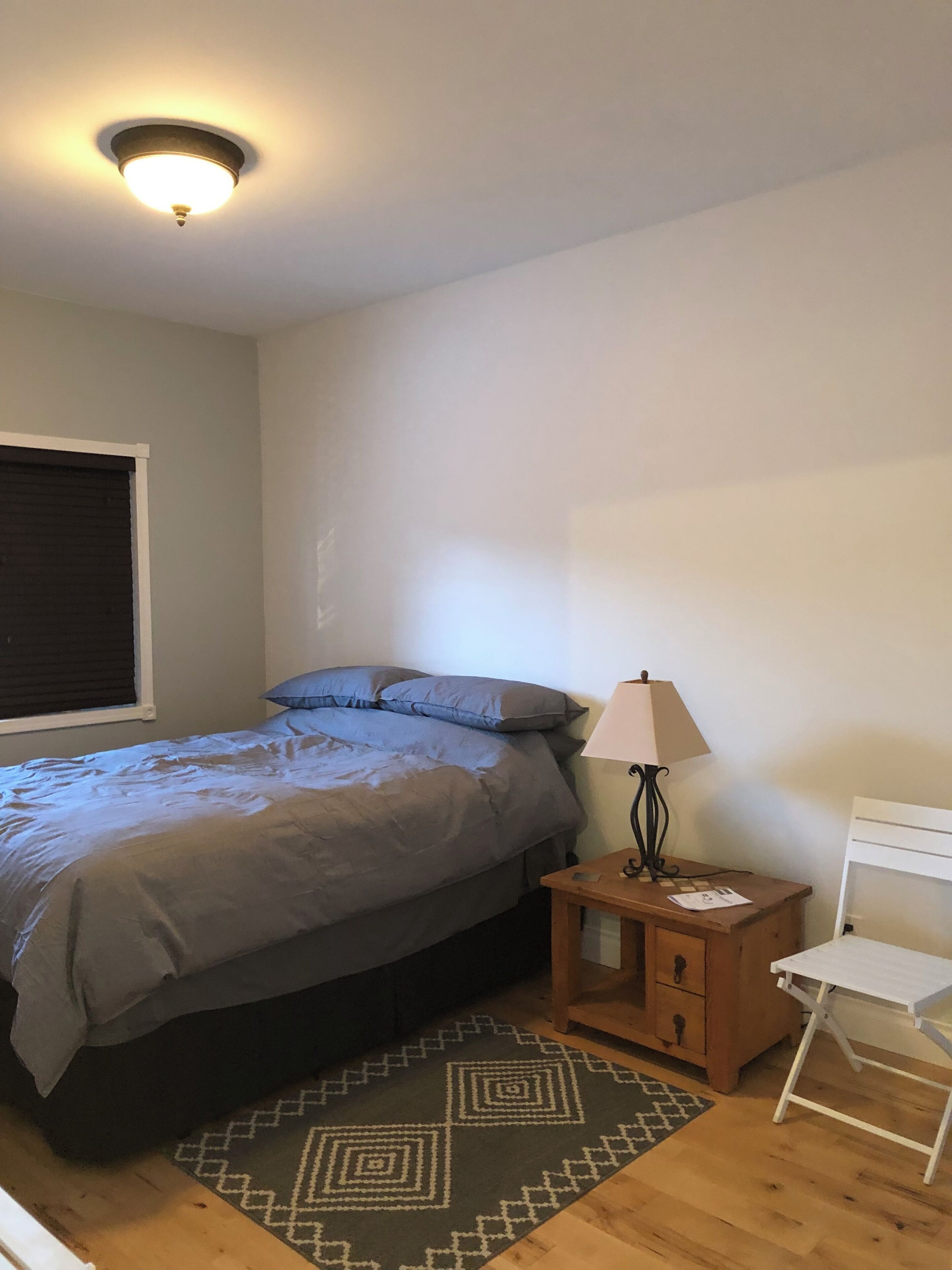 Een slaapkamer met een bed, nachtkastje, lamp, stoel en een kamerkleed met een patroon.