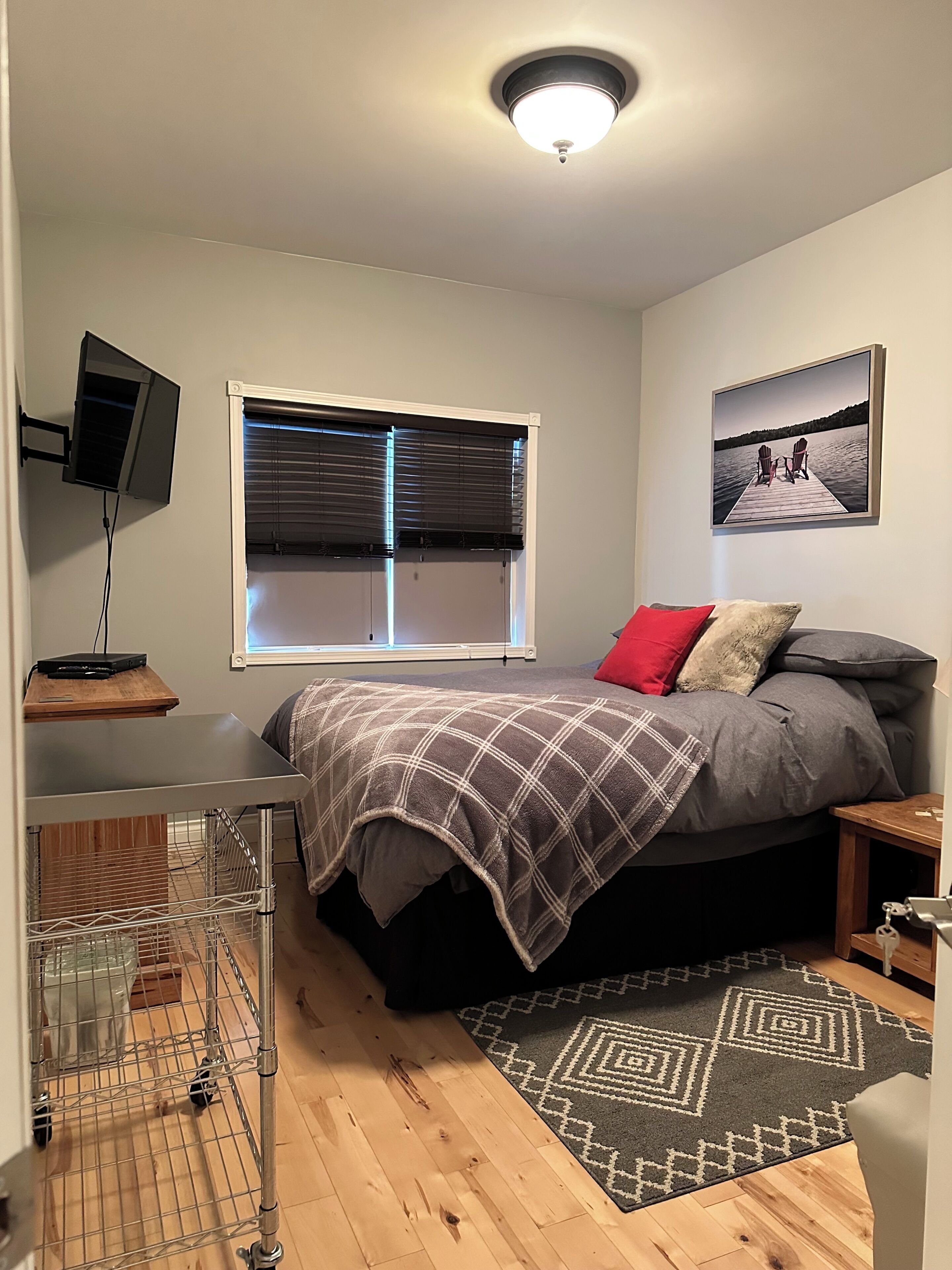 Une chambre à coucher avec un lit, une fenêtre avec des stores, un téléviseur fixé au mur, un tableau au mur et une petite table.